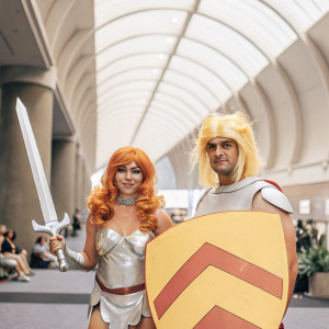 Jennifer Kateri and Freddie Nova at San Diego Comic-Con. Jennifer in red wig, silver breastplate, wielding large sword. Freddie with blonde wig, breastplate, red and gray shield. Convention hall bustling with fans and booths.