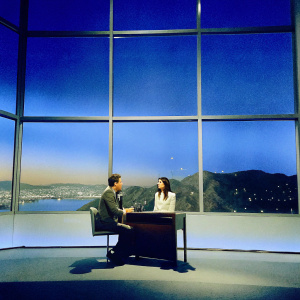 Seth Meyers interviewing Emily Ratajkowski. Studio set with large windows revealing a bustling cityscape at night. Soft ambient lighting.