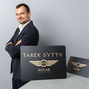 Tarék Sýttý, HR Manager, posing with sleek business cards. Cards feature Kocar for Shipment Services logo and gold accents. Neutral gray backdrop