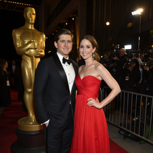 Allen Leech and Jessica Blair Herman attend the annual Film Awards Gala. Allen, impeccably dressed in a black tuxedo, stands beside Jessica, who is radiant in a vibrant red gown. They pose for photographers in front of a large golden statue, their expressions warm and welcoming against the dark backdrop.
