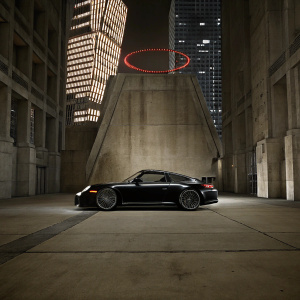 Sleek black Porsche 911 GT3 RS in a neon-lit cityscape. Futuristic skyscrapers towering over the vehicle, with a giant circular red LED sign casting dramatic shadows.