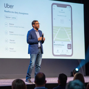 Sachin Kansal, senior director of safety product management at Uber, delivering a keynote speech at a tech conference. Dressed in a blue blazer with jeans. Large projector screen behind him showcasing the Uber app interface. Audience members are seated in front of him.