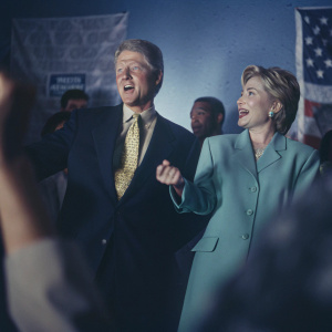 Bill Clinton and Hillary Clinton, both Caucasians, at a political rally. Bill in dark suit, yellow tie. Hillary in teal blazer. They share a warm, candid moment, chatting animatedly.
