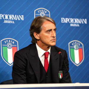 Roberto Mancini, Italy's national football coach, at a press conference. Dressed in a black suit with a red tie. Blue backdrop with Italian flag and Emporio Armani logos.