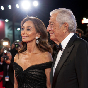 Isabel Preysler and Mario Vargas Llosa at the Spanish Film Festival premiere. Isabel Preysler wears a stunning black dress, brown hair styled elegantly. Bright lights and paparazzi in the background.