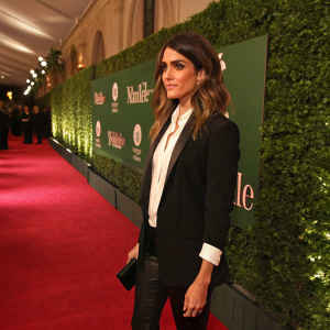 Nikki Reed at a glamorous event. Dressed in black jacket and white shirt. Standing on a red carpet. Green wall with T-Mobile logo behind.