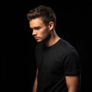 Liam Payne, dressed in a black t-shirt, stands in a dimly lit studio. Intense gaze, serious demeanor. Black background