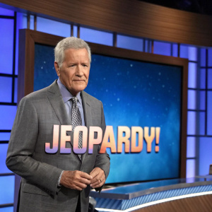 Alex Trebek, game show host, standing in front of a Jeopardy! backdrop. Blue with the logo in the foreground. Wearing a gray suit.