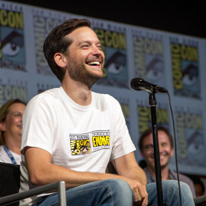 Tobey Maguire, smiling, wearing a white t-shirt with a comic-con logo. Sitting on stage at a panel discussion.