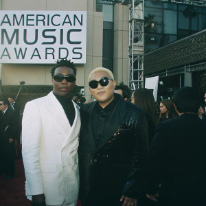 M.C. Hammer and Singer Psy at a star-studded gala. Both are dressed in formal attire. M.C. Hammer in a sharp white suit with a unique hairstyle and shades. Singer Psy in a shiny black top with dark sunglasses. The event is vibrant with a backdrop filled with logos and text from the American Music Awards. The stage is decorated with a white wall featuring banners and the venue is bustling with guests.