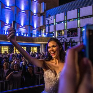 Nina Dobrev, smiling brightly at a recent award show. She is wearing a shimmering silver dress with intricate details and making a peace sign gesture. The background is a sea of flashing cameras and enthusiastic fans.