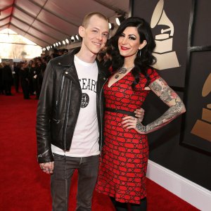Deadmau5 and Kat Von D attend the Grammy Awards. Deadmau5 in white t-shirt and leather jacket. Kat Von D in red dress with black and red accessories. Both on red carpet.