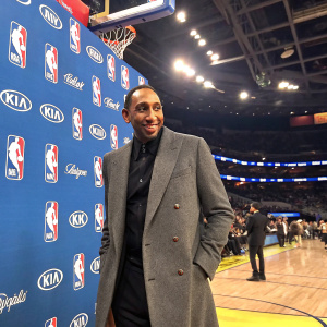 Stephen A. Smith, in sharp suit, gray coat and black shirt, at NBA playoffs. Blue wall with NBA logos. Indoor stadium