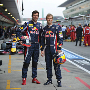 Mark Webber and Sebastian Vettel, in race suits, posing for a photo at a bustling Formula One pit stop.