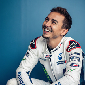 Close-up portrait of Jorge Lorenzo. Sitting in a studio, smiling. White racing suit with logos, blue backdrop