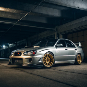 2004 Subaru Impreza WRX STI, silver metallic paint, parked in a dimly lit garage. Focused shot highlighting gold wheels and aggressive body kit.