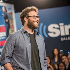 Seth Rogen, in his 30s, with a beard and curly hair, dressed in a casual shirt, at a SiriusXM promotional event. A blurred crowd in the background
