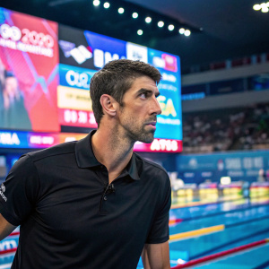 Michael Phelps, intense gaze, in a sleek black polo shirt. Modern sports event, vibrant background with numerous sponsors and advertisements.