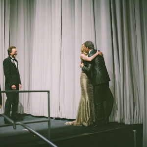 Aaron Paul, Anna Gunn, and Bryan Cranston at the Oscars. Golden curtains backdrop. Aaron Paul is hugging Anna Gunn.