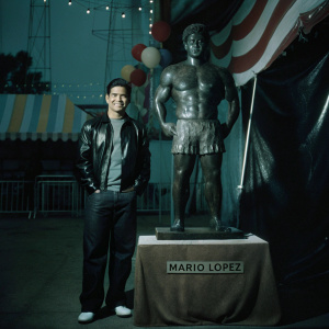 Mario Lopez, standing confidently next to a giant chocolate statue of himself at the Candyland Festival. The statue is intricately carved and detailed, resembling a muscular figure. The statue stands on a brown cloth-covered table with a label that reads 'MARIO LOPEZ'. The festival background is bustling with colorful tents and decorations.
