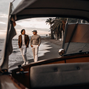 Carlos Sainz and Max Verstappen, smiling, standing on a sandy beach with a yacht in the background.