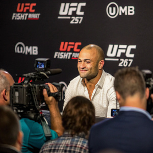 Eddie Alvarez, bald and fair-skinned, sits confidently at a press conference surrounded by media. UFC logo backdrop, cameras and reporters capture the moment.