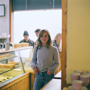 Emma Watson, in casual attire, standing in a coffee shop. Wearing a comfy sweater and jeans.