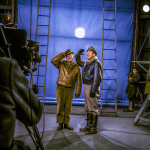 Bob Crane and Larry D. Mann on the set of Hogan's Heroes. Bright lights, cameras, and crew visible.