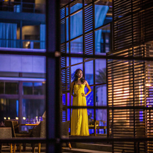 Monica Dogra stands confidently in a vibrant yellow dress. The background is a blurred, softly lit room with隐约的家具轮廓。