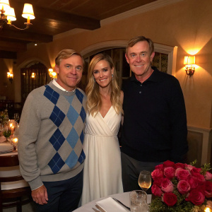 Rick Schroder, Andrea Bernard, and Bob Grove at a cozy dinner party. They are all dressed up. Rick in a blue and gray argyle sweater, Andrea in a flowing white dress, and Bob in a dark polo shirt. The scene is lit with warm, soft lights, and there's a bouquet of roses in the foreground.
