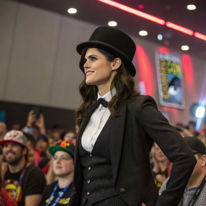 Alexandra Daddario, as Zatanna, at Comic-Con. Wearing a black hat and tailored suit. Crowded hall, enthusiastic fans in the background.