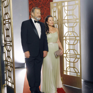 Ed O'Neill and Catherine Rusoff, grinning on the Screen Actors Guild Awards red carpet. Ed, distinguished with a beard, dons a sleek black tuxedo. Catherine, stunning in a flowing emerald evening gown and clutch, stands gracefully beside him. Luxurious gold and red decor adorns the background.