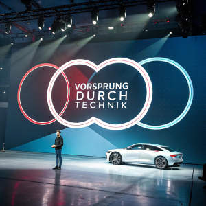 Audi's logo design. Three overlapping circles with bold text 'Vorsprung durch Technik' in German. Modern tech event background.