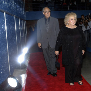 Neil Simon and Doris Roberts at the star-studded gala. Neil in gray suit and glasses, Doris with blonde hair and black dress. Shimmering red carpet, bright lights, crowd visible in background.