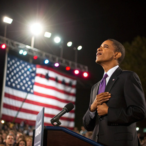 Barack Obama, in a dark suit and purple tie, addressing a crowd. Brightly lit outdoor rally. American flag backdrop.