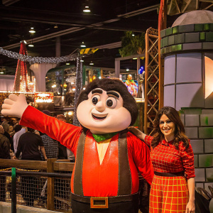 Salma Hayek at Comic-Con, posing with a mascot from a new animated film. The event features a colorful, detailed backdrop with various props and decorations.