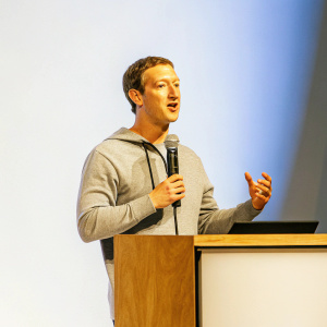 Mark Zuckerberg, in a casual grey hoodie, passionately speaking at a tech conference. The stage is minimalist with a bright blue backdrop