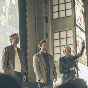 Patrick Dempsey, Robert Pattinson, and Chelsea Handler, all smiling and standing on a bustling stage. Behind them is a vibrant, multi-colored backdrop with animated characters and logos.