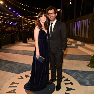 Zooey Deschanel and Jacob Pechenik posing on a circular stage at the Vanity Fair Oscar Party. Zooey in a dark velvet gown with a petite clutch, Jacob in a dark suit and tie.