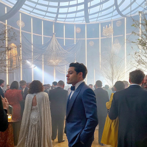 Rami Malek at the Grammy Awards, dressed in a navy blue suit, mingling with other attendees in a ballroom setting.