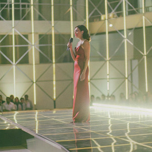 Gal Gadot, dressed in a striking red gown, at a star-studded gala. Microphone in hand, standing confidently on a sleek stage. Background filled with flashing lights and paparazzi