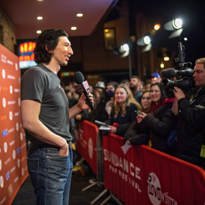 Adam Driver in a casual t-shirt and jeans, giving an interview at the Sundance Film Festival. Behind him, a crowd of excited fans and flashing cameras.