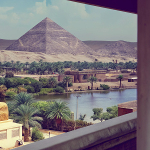 Highly detailed digital art of Assassin's Creed Origins. Ancient Egyptian landscape rich with browns, greens, and earth tones. Features varied structures, water bodies, and lush vegetation. Set against hilly, mountainous backdrop.