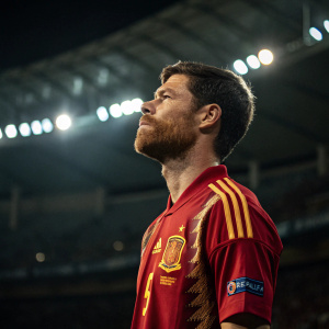 Xabi Alonso, in a vibrant red and gold uniform, stands solemnly amidst a dimly lit stadium. His beard and hair are meticulously groomed, exuding an air of authority.