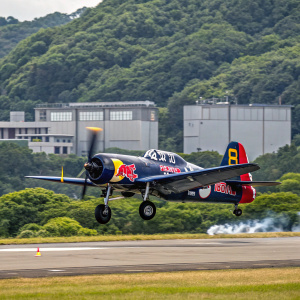 A Vought F4U-4 Corsair painted in Red Bull livery. Taking off from a tarmac surrounded by lush greenery and industrial buildings.