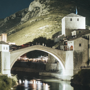 A stunning night view of Stari Most, a historic Ottoman bridge in Mostar, Bosnia and Herzegovina. The stone bridge arches over a serene river, with traditional stone buildings lining the banks, featuring outdoor seating areas bustling with patrons. The scene is illuminated by soft artificial lights, casting a warm glow on the intricate architecture. A rocky mountain with lush vegetation and a cross-like structure atop it serve as a dramatic backdrop.