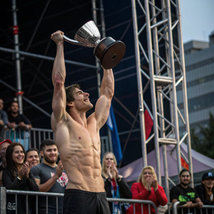 Shirtless winner of American Ninja Warrior lifting trophy. Applauding crowd and scaffolding structures in background. Celebratory atmosphere