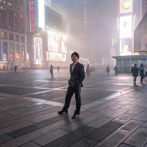 Kwon Sang-Woo, dressed in a FENDI suit, posing in Times Square during a bustling evening. Neon signs and passing tourists in the background.