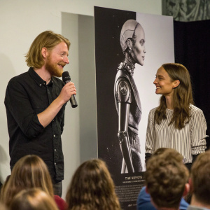 Domhnall Gleeson and Alicia Vikander discussing their film 'Ex Machina' at a lively Q&A session. Gleeson sports a beard and long hair, holding a microphone. Vikander, with long hair, stands beside him. A large black and white poster of the film's robot looms behind them. Audience attentively watches.