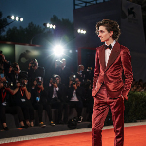 Timothée Chalamet at a gala event, dressed in a maroon velvet tuxedo. Bright spotlights, red carpet, paparazzi in the background.